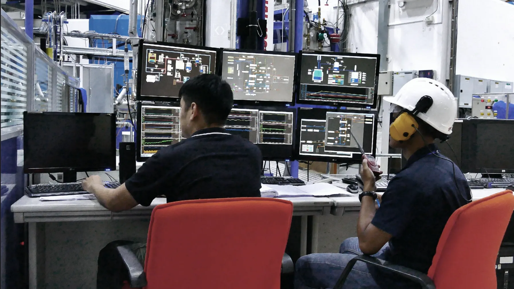 Two operators at an industrial control room workstation surrounded by monitoring screens