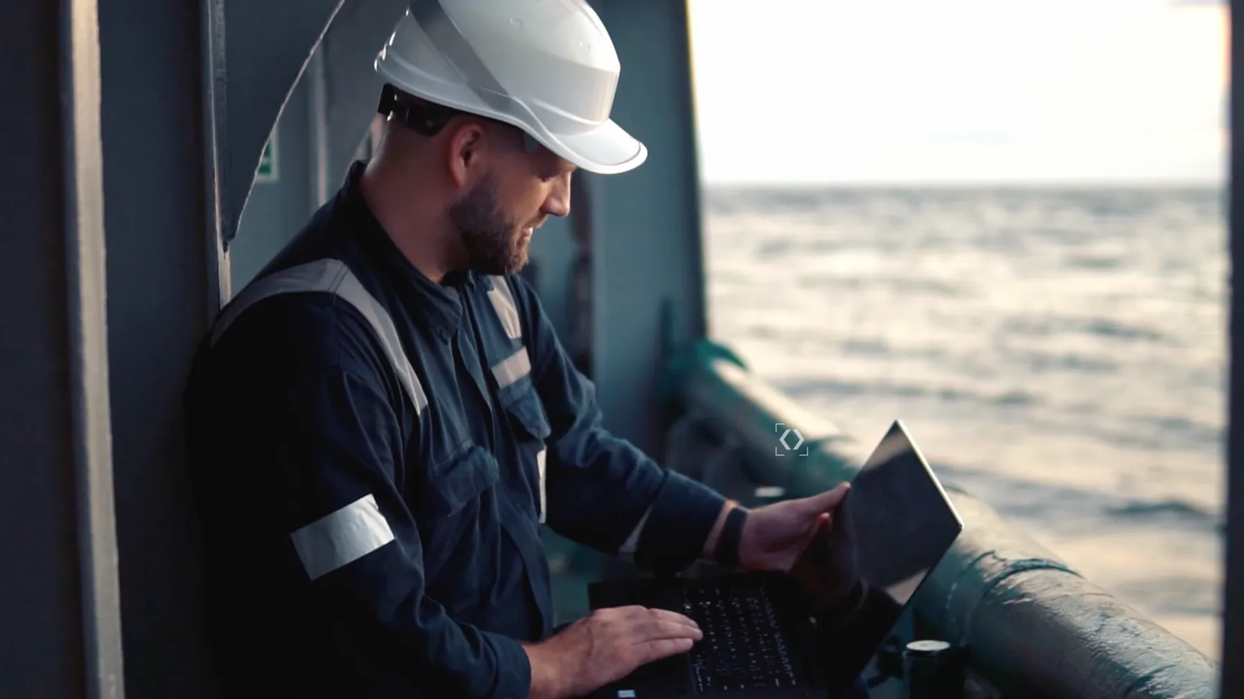 An engineer in a hard hat working on a laptop on the deck of a ship at sea